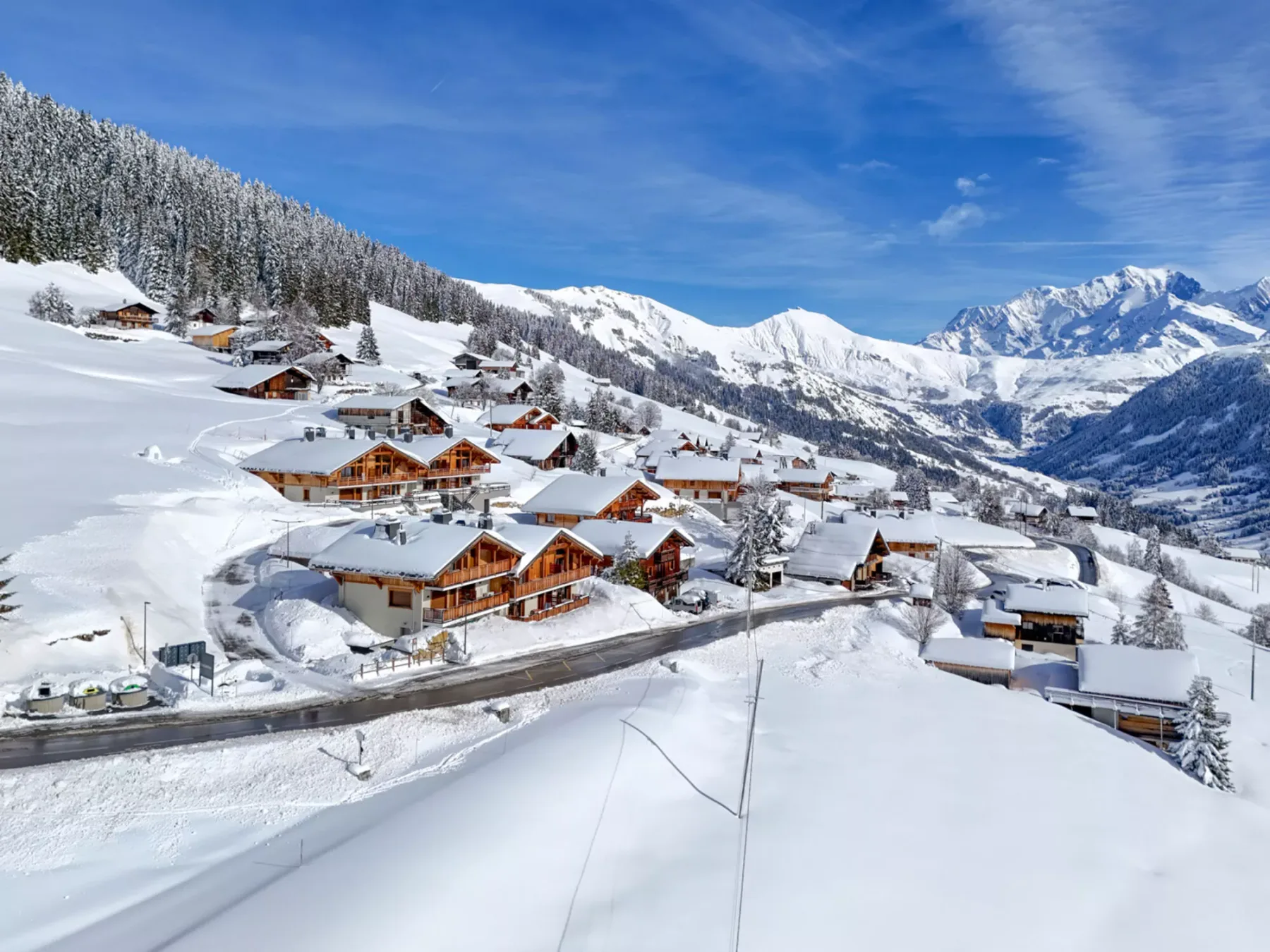 A snow-covered alpine village sits below forested slopes and distant mountain peaks.