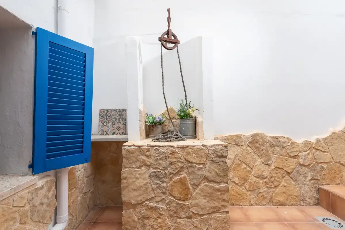 A stone well stands in a small courtyard with white walls and blue shutters.