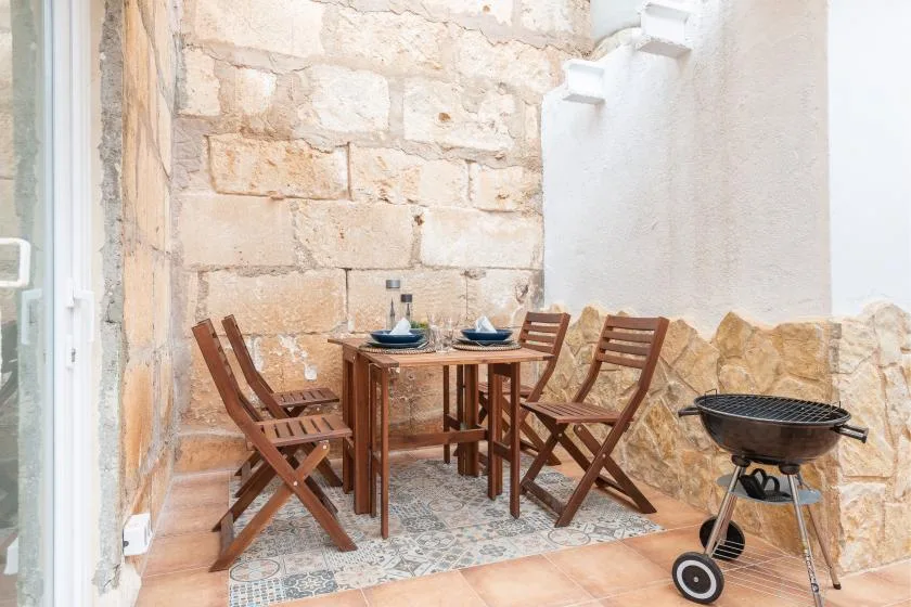 A small patio dining area has a wooden table and chairs beside a portable grill.