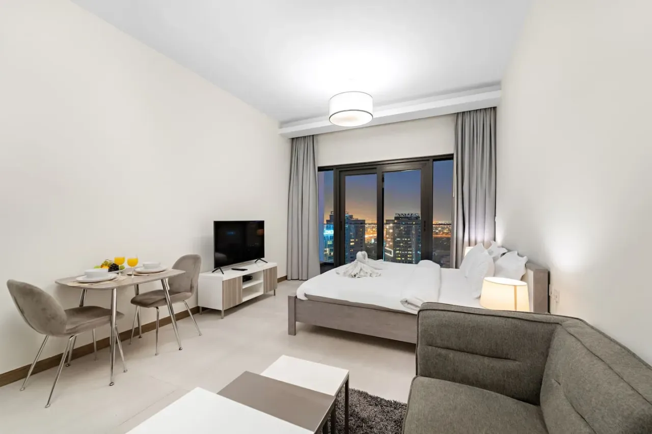 A bedroom with a bed, dining table, and city views through large windows.