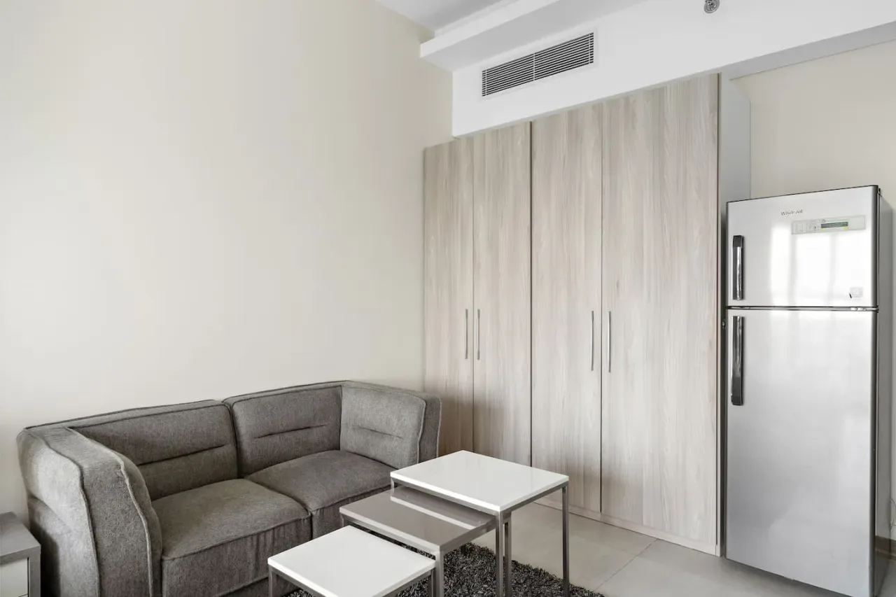 A living area with gray seating, a small table, and a refrigerator beside tall cabinets.