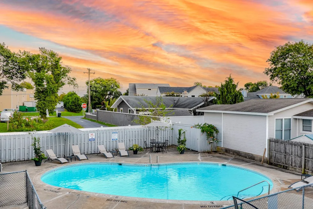 An outdoor swimming pool sits beside a white fence under a colorful sunset sky.