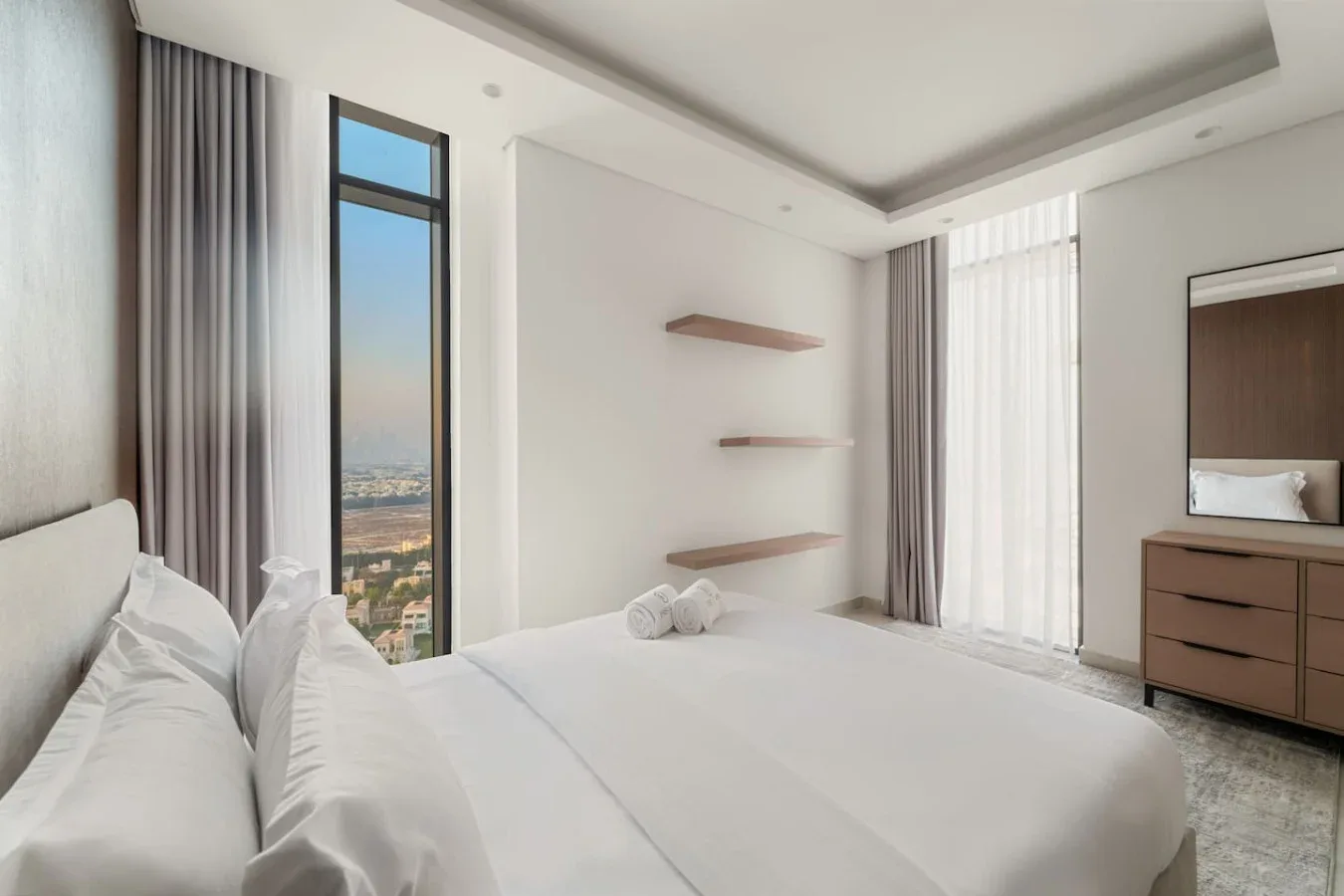 A bright bedroom with white bedding, tall windows, and floating wall shelves.