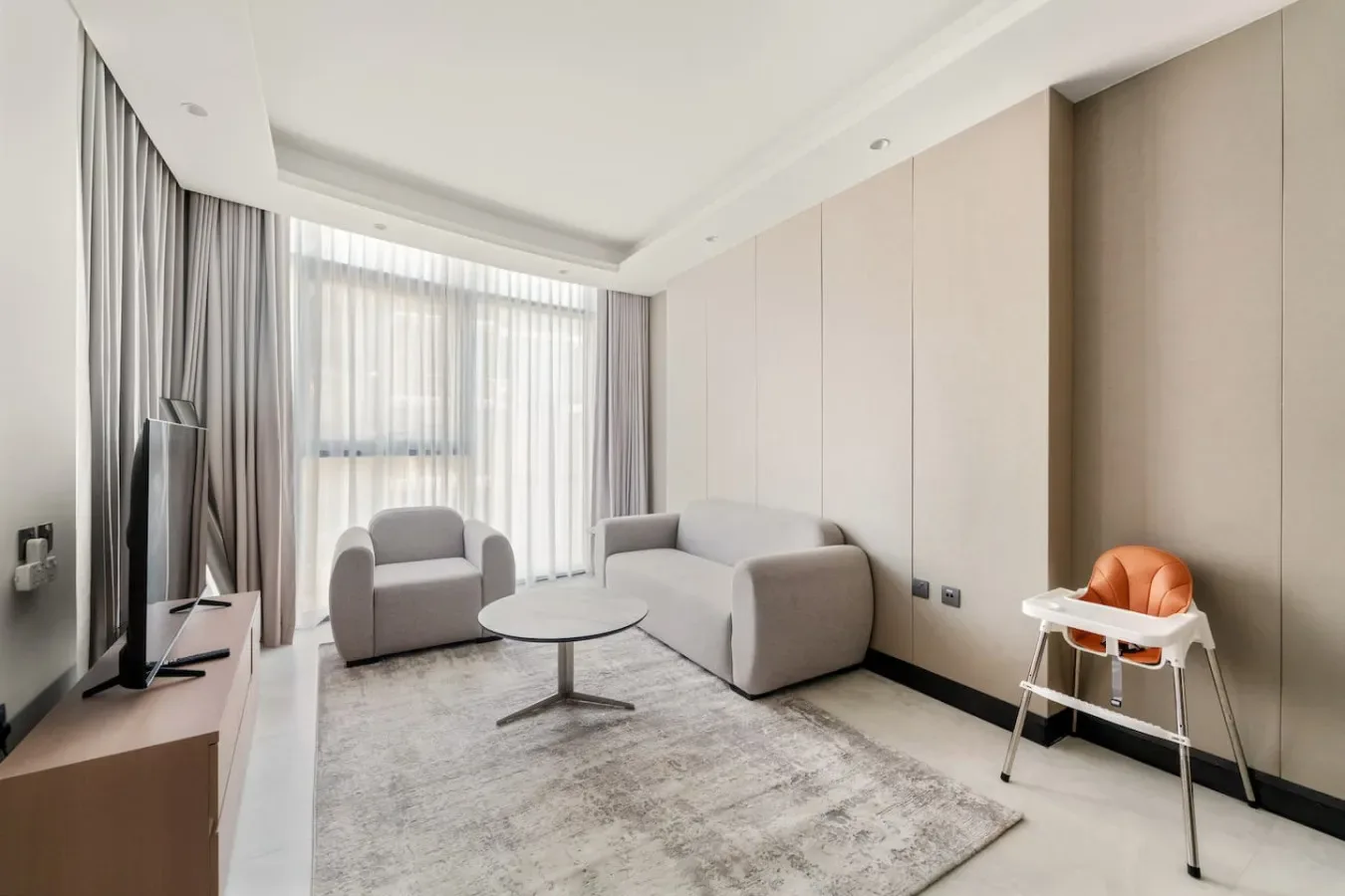 A bright living room with seating, a television, and large curtained windows.