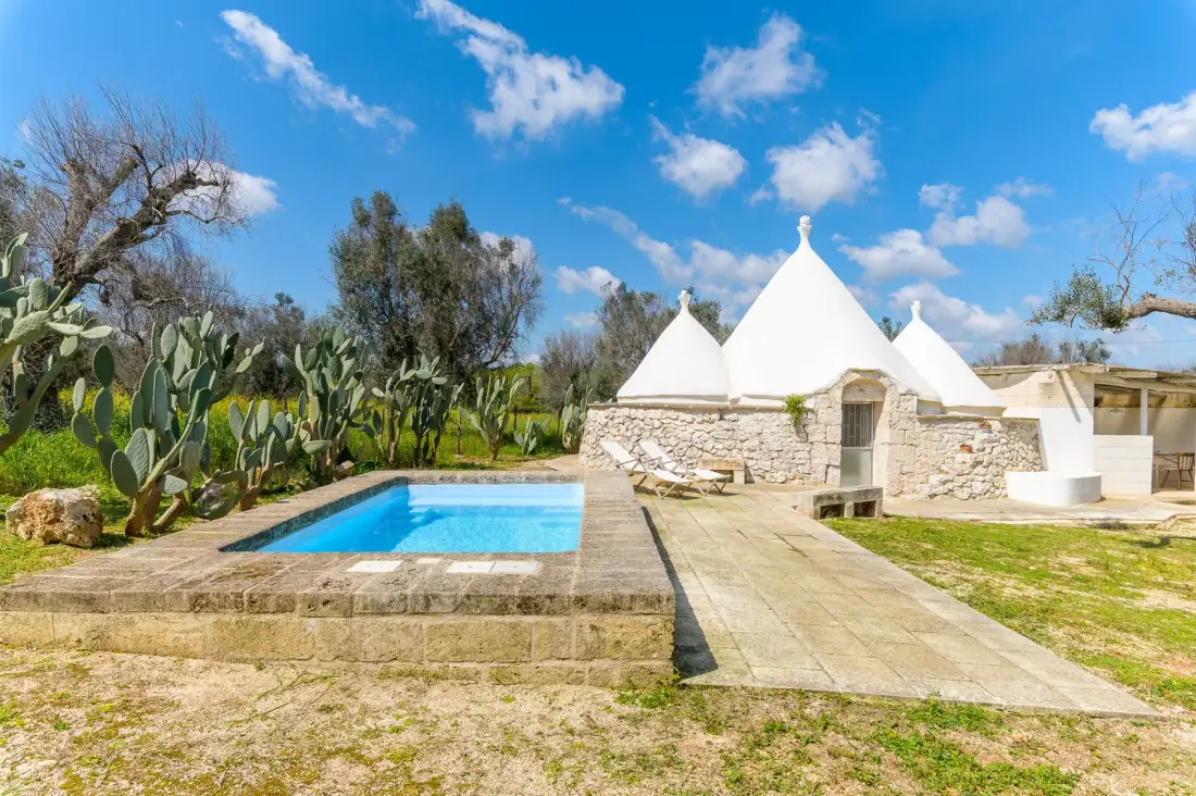 An outdoor swimming pool sits beside white conical-roofed stone buildings.