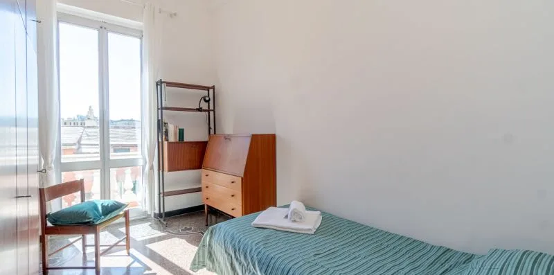 A bedroom with a bed, wooden desk, and tall windows opening onto a balcony.