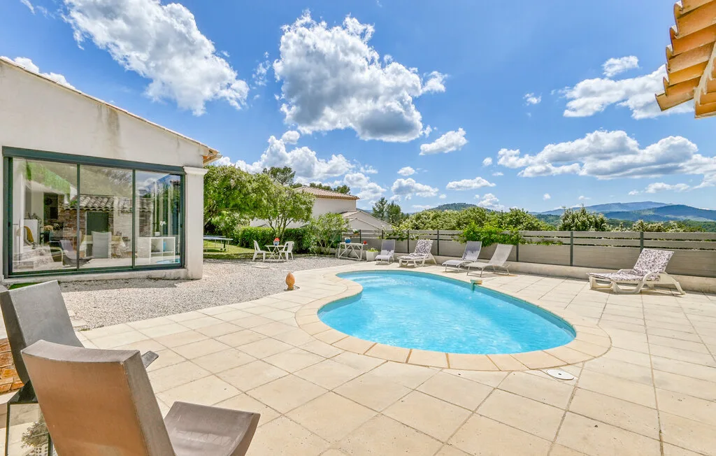 An outdoor pool sits in a spacious paved patio beneath a bright sky.