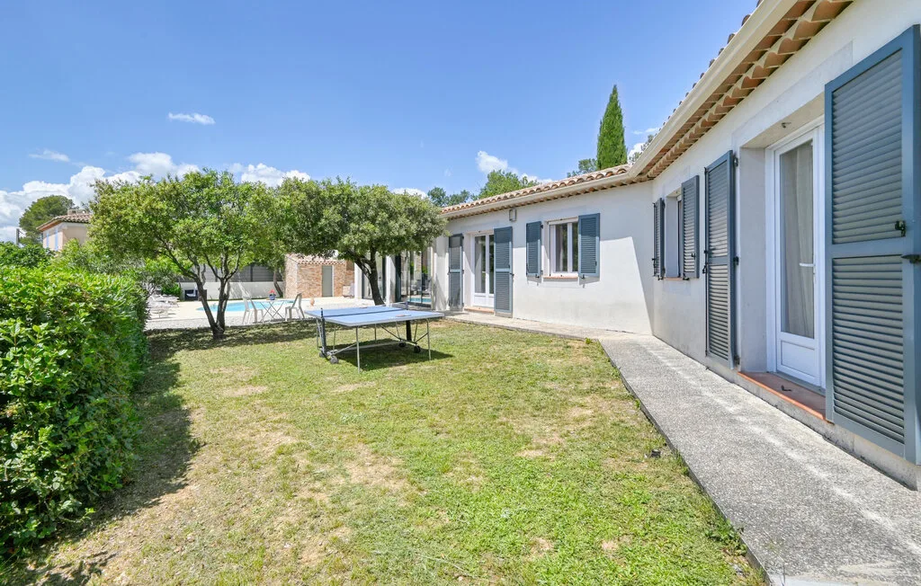A sunny garden patio with a lawn, leafy trees, and a paved walkway beside the house.