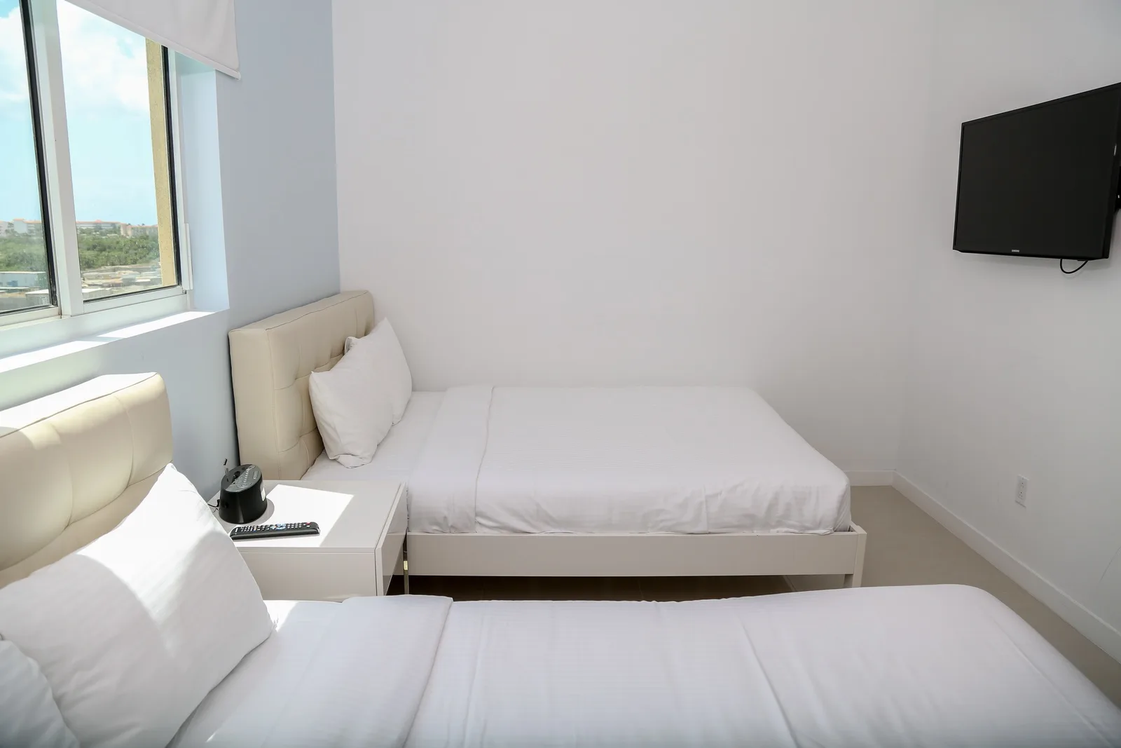 Your bright, modern bedroom awaits with two crisp white beds, a mounted TV for your entertainment, and beautiful natural light streaming through the window offering peaceful views.