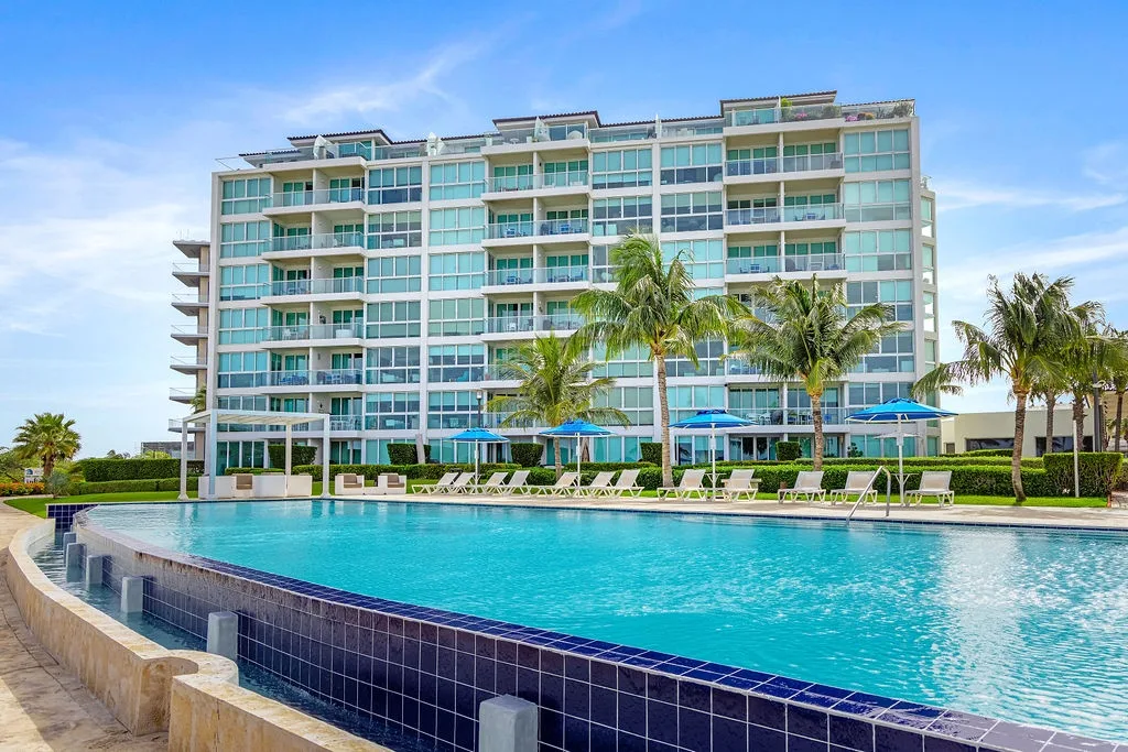 Modern beachfront resort building with pristine pool area surrounded by swaying palms under sunny skies.