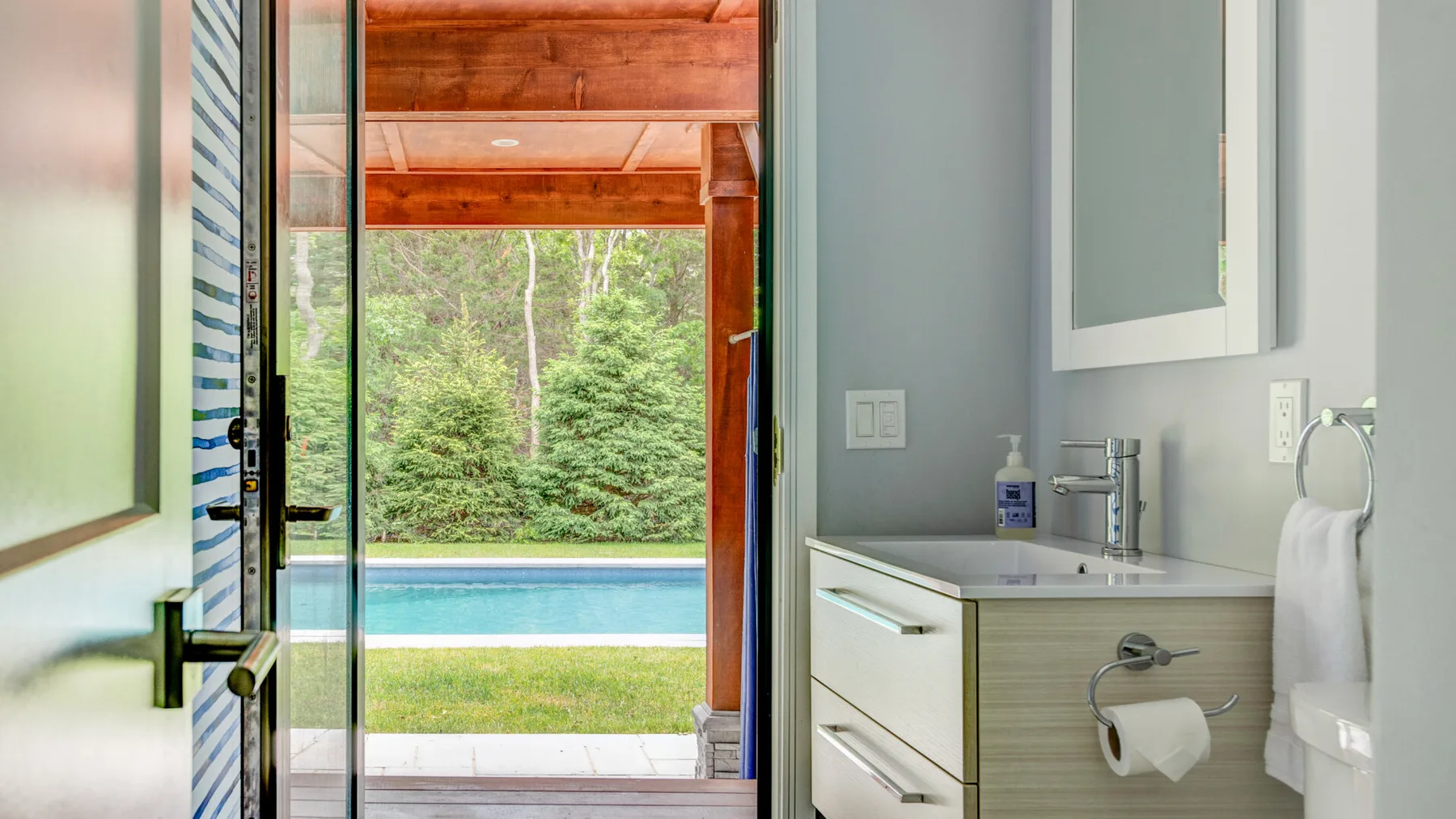 A bathroom vanity and sink overlook a glass door opening to greenery and a pool.