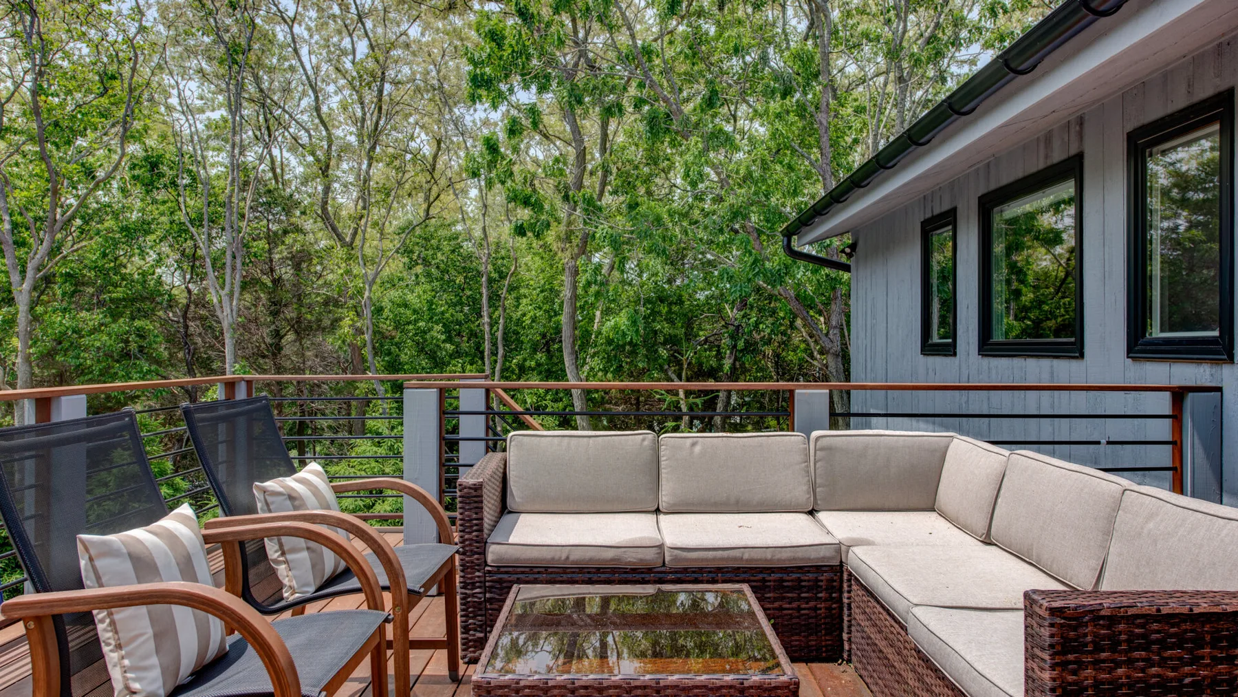 An outdoor deck has an L-shaped seating area beside a wooded backdrop.