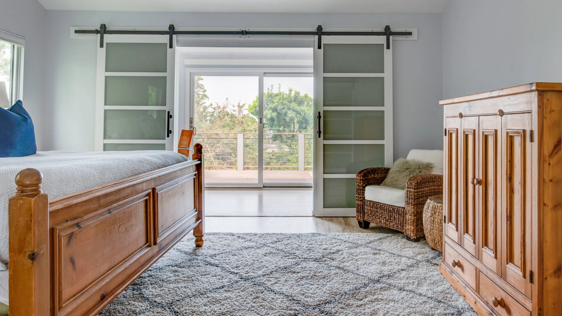 A bright bedroom with a wooden bed, sliding glass doors, and a woven armchair.