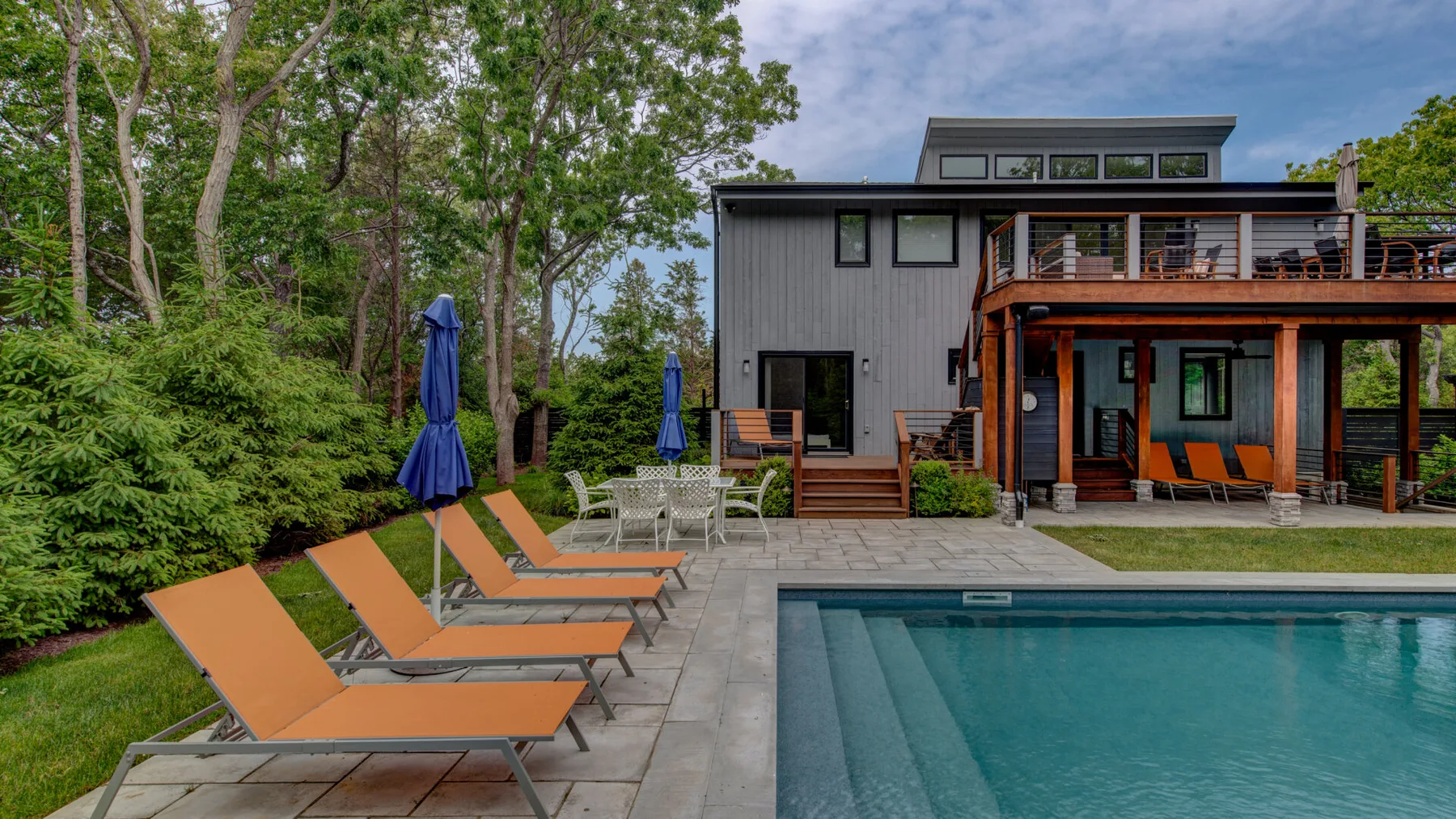 A swimming pool and lounge seating sit beside a modern house, surrounded by trees.