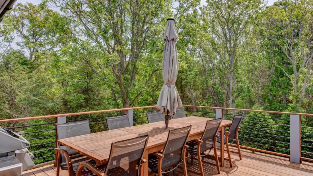 An outdoor dining area with a wooden table and chairs overlooks leafy trees.