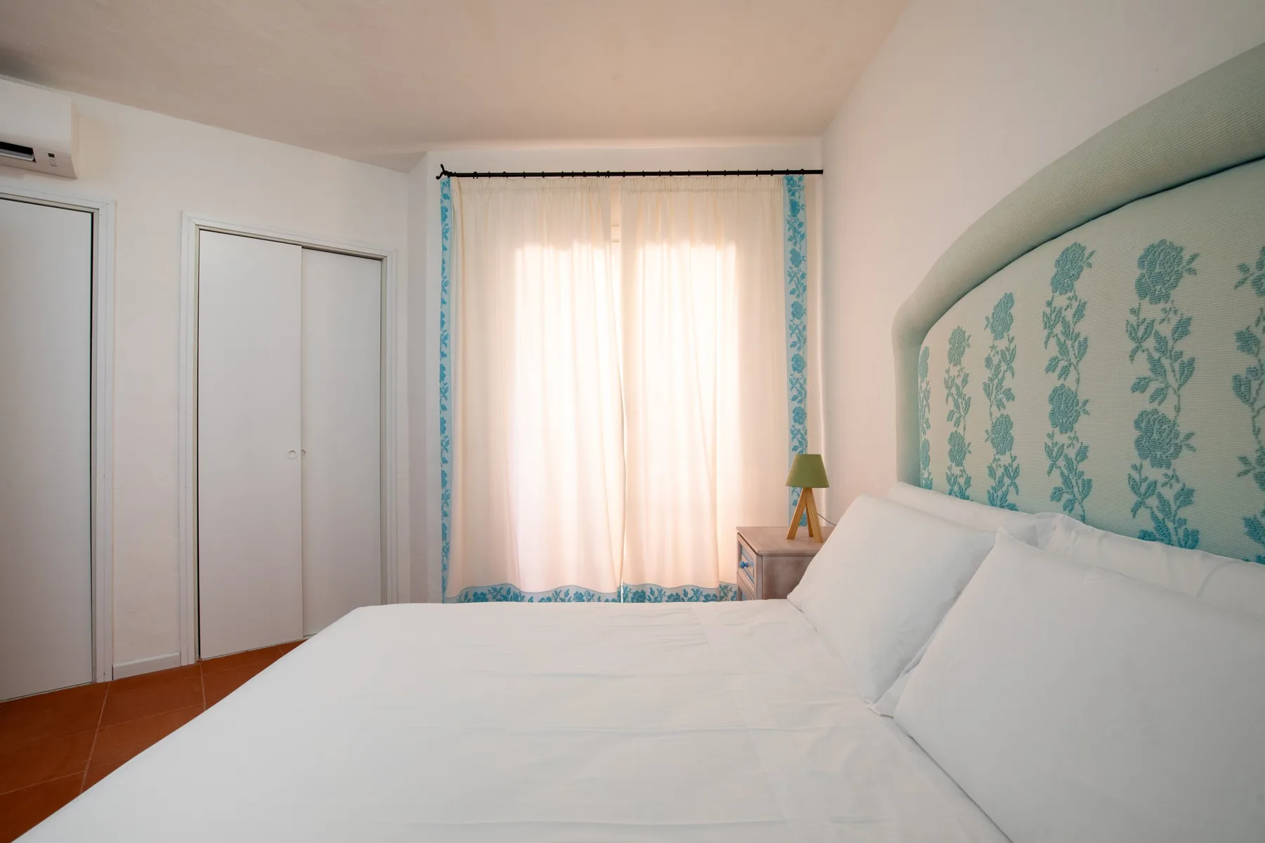 A bedroom with a large bed, floral wall covering, and a bright window.