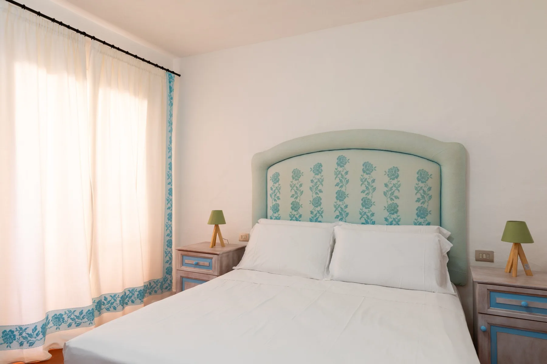 A bedroom with a white bed, patterned headboard, and sheer curtains.