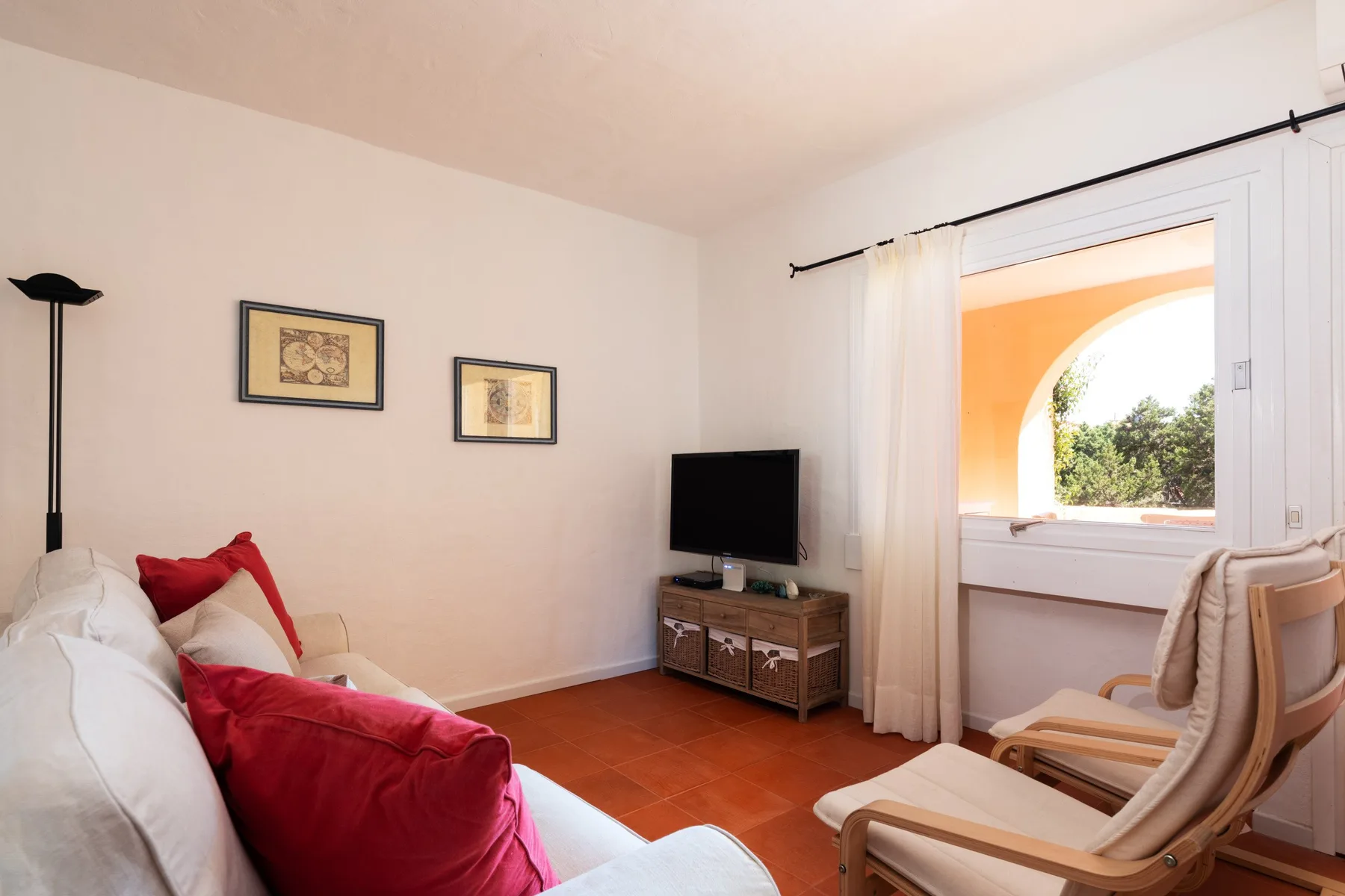 A bright living room with red cushions and a television beside an arched window.