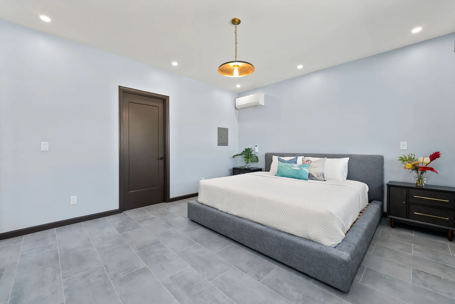 A bright bedroom with a bed, dark door, and tiled floor.