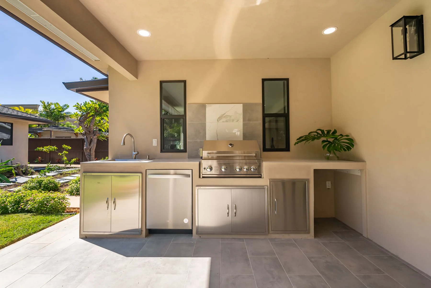 An outdoor kitchen with a grill, sink, and counter beneath a covered patio.