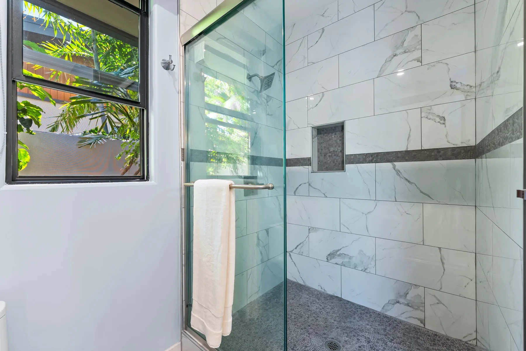 A tiled shower with a glass panel sits beside a window overlooking greenery.