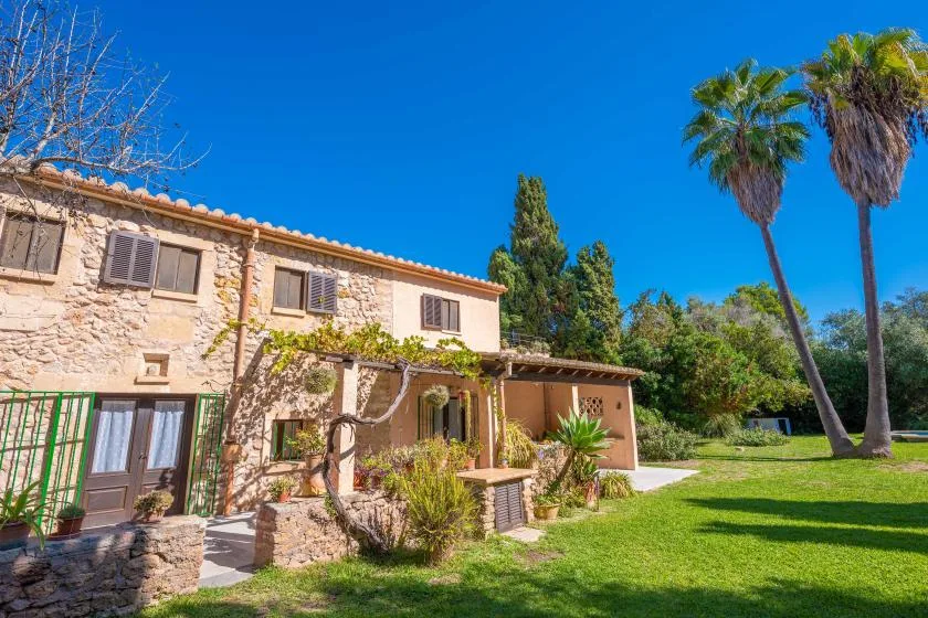 A stone house opens onto a sunny garden with lawn and palm trees.