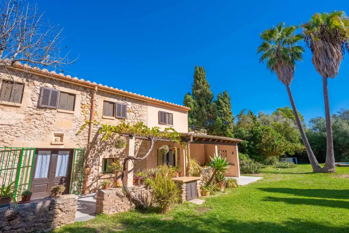 A stone house stands beside a green lawn and several palm trees.