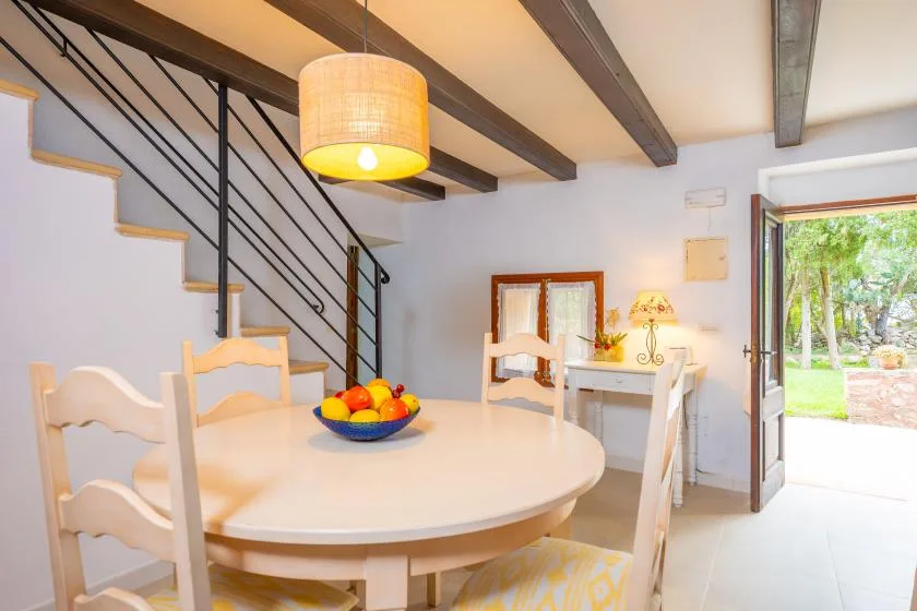 A dining area with a round table sits beneath exposed ceiling beams beside a staircase.