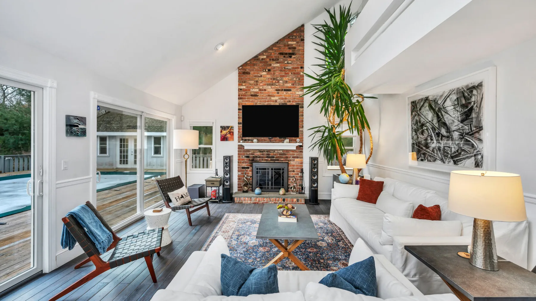 A bright living room features a brick fireplace, seating, and large windows overlooking a pool.
