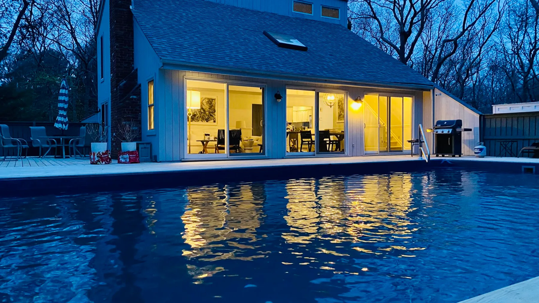 A swimming pool reflects the warm lights of a house at dusk.