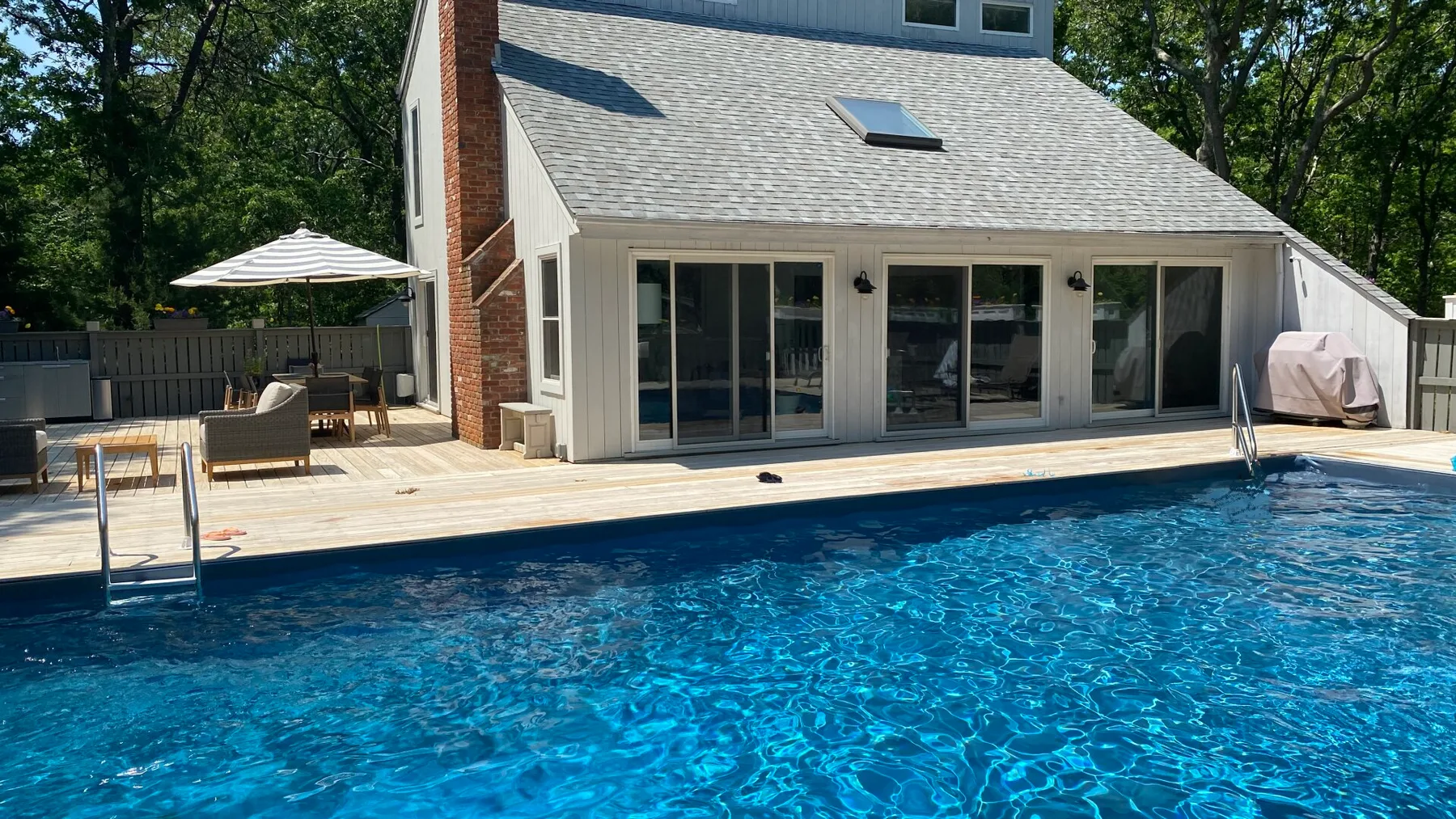 A swimming pool beside a house, with a wooden deck and patio seating.