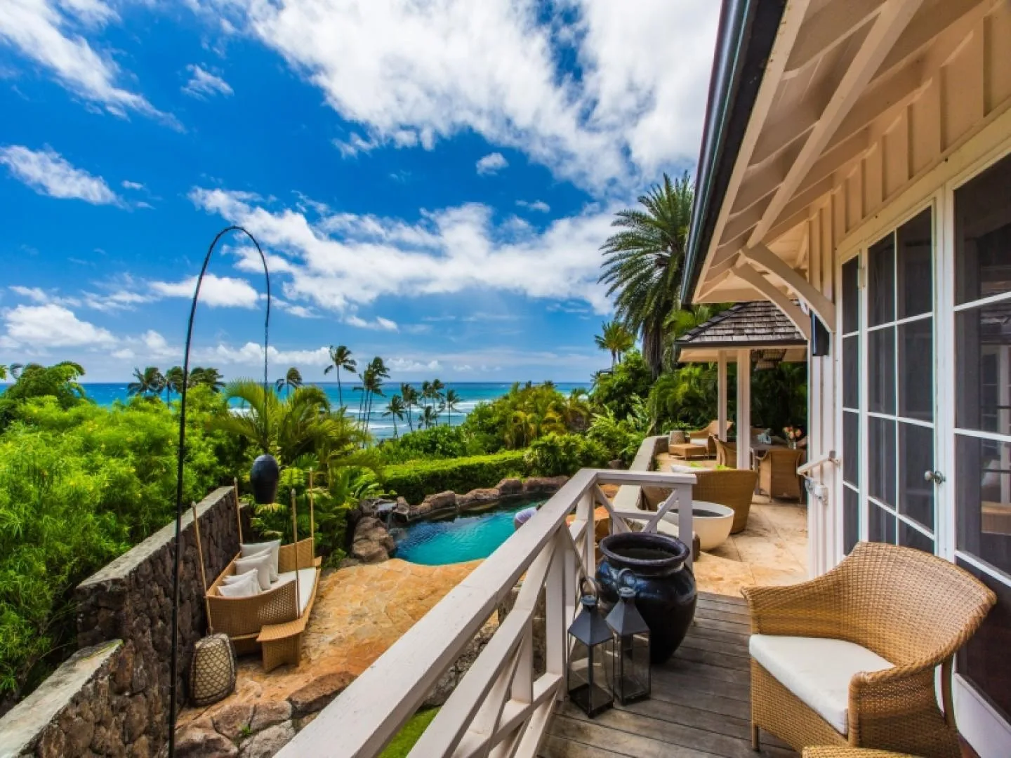 A furnished outdoor terrace overlooks a pool, lush greenery, and the ocean.