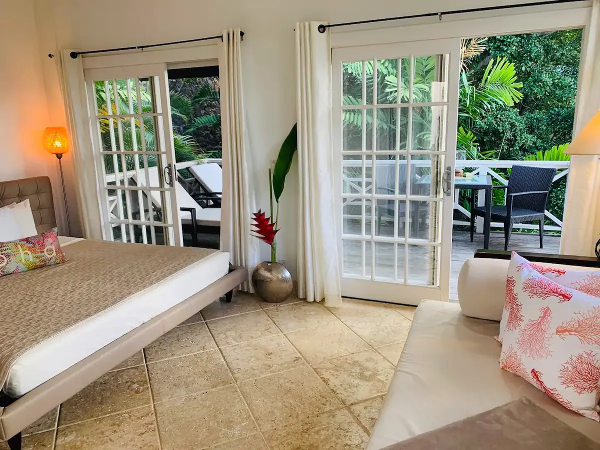 A bedroom with a bed and seating beside glass doors opening onto a leafy patio.