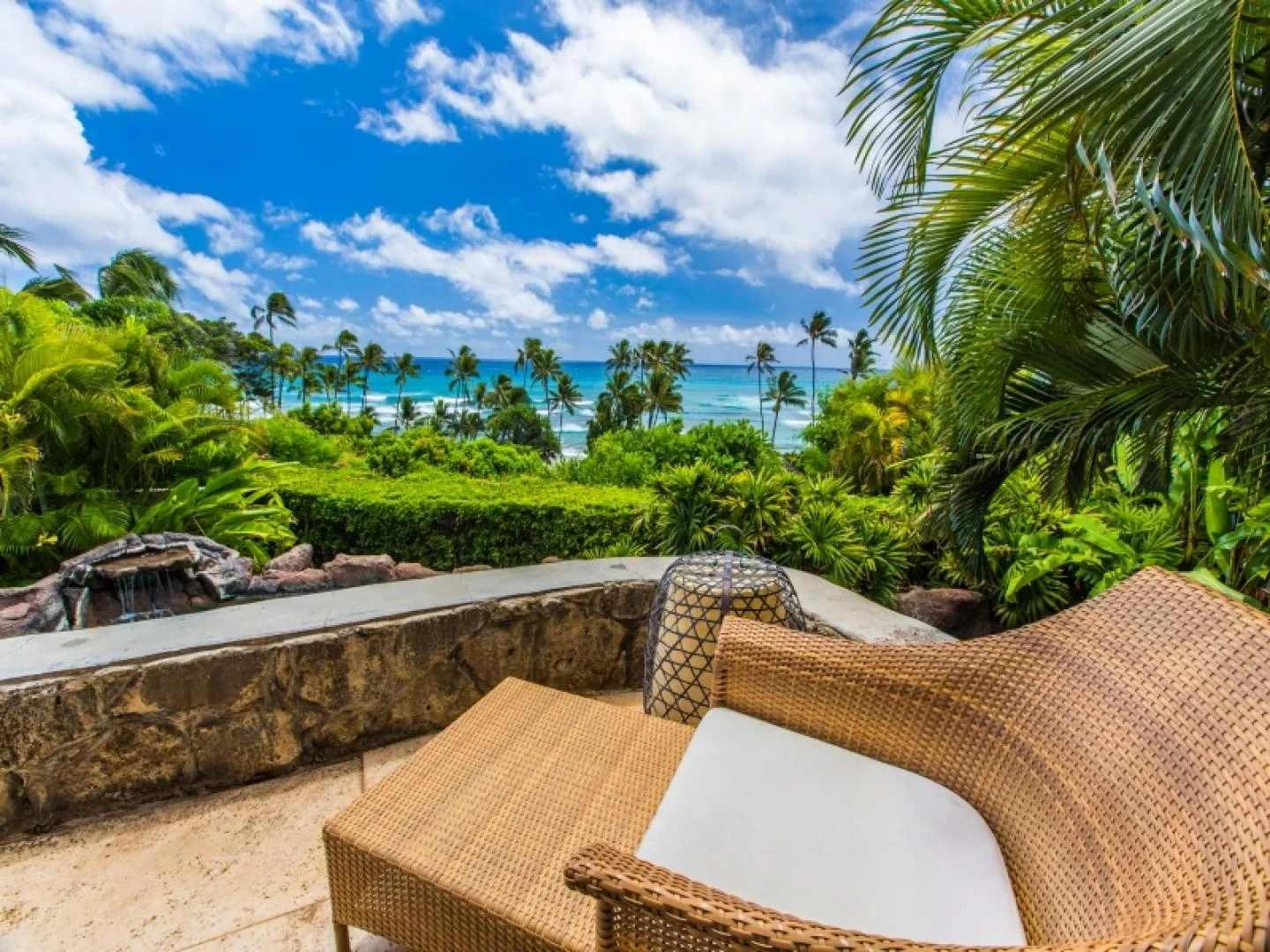 A woven lounge chair sits on a terrace overlooking lush tropical vegetation and the ocean.