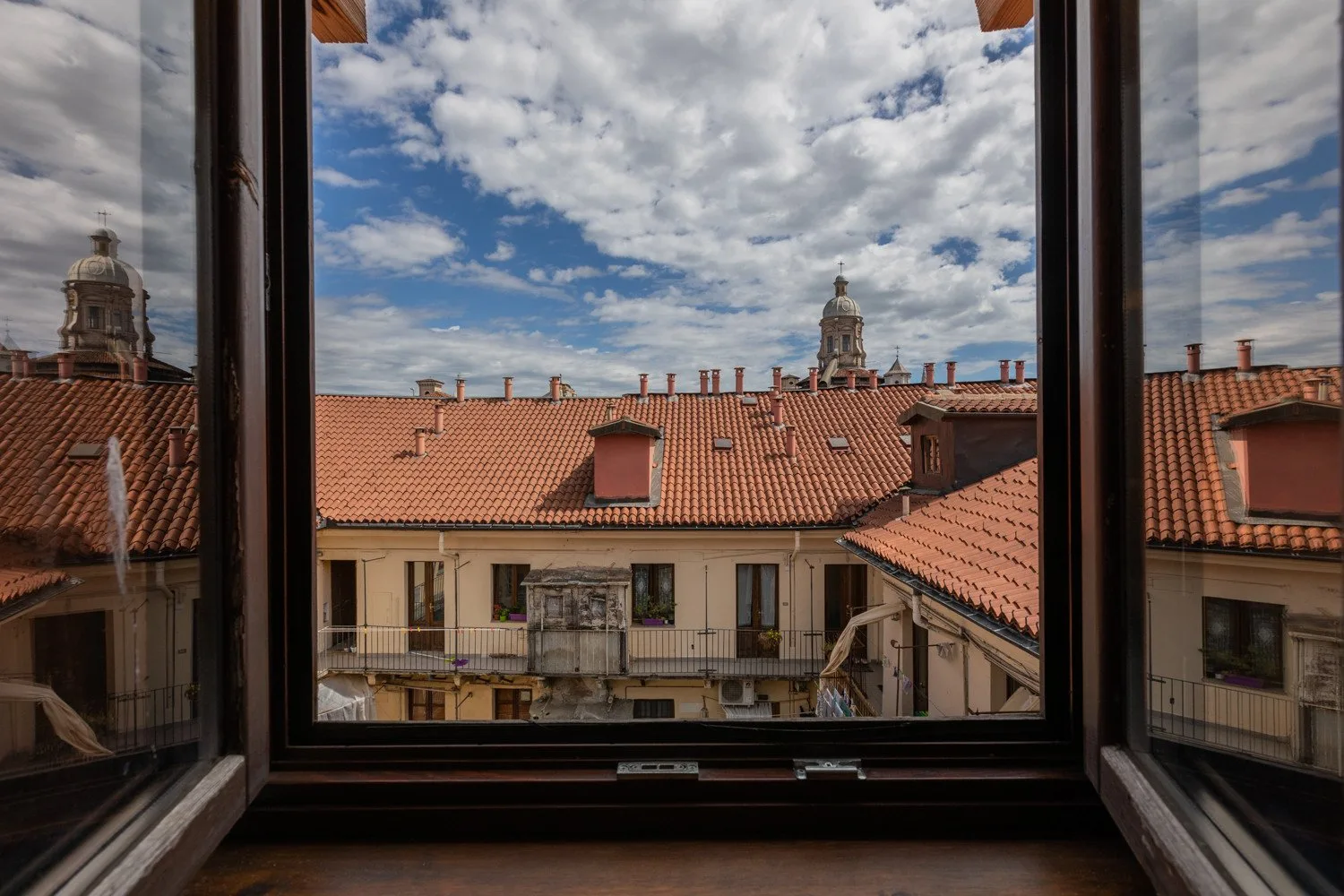 An open window overlooks red-tiled rooftops beneath a cloudy blue sky.