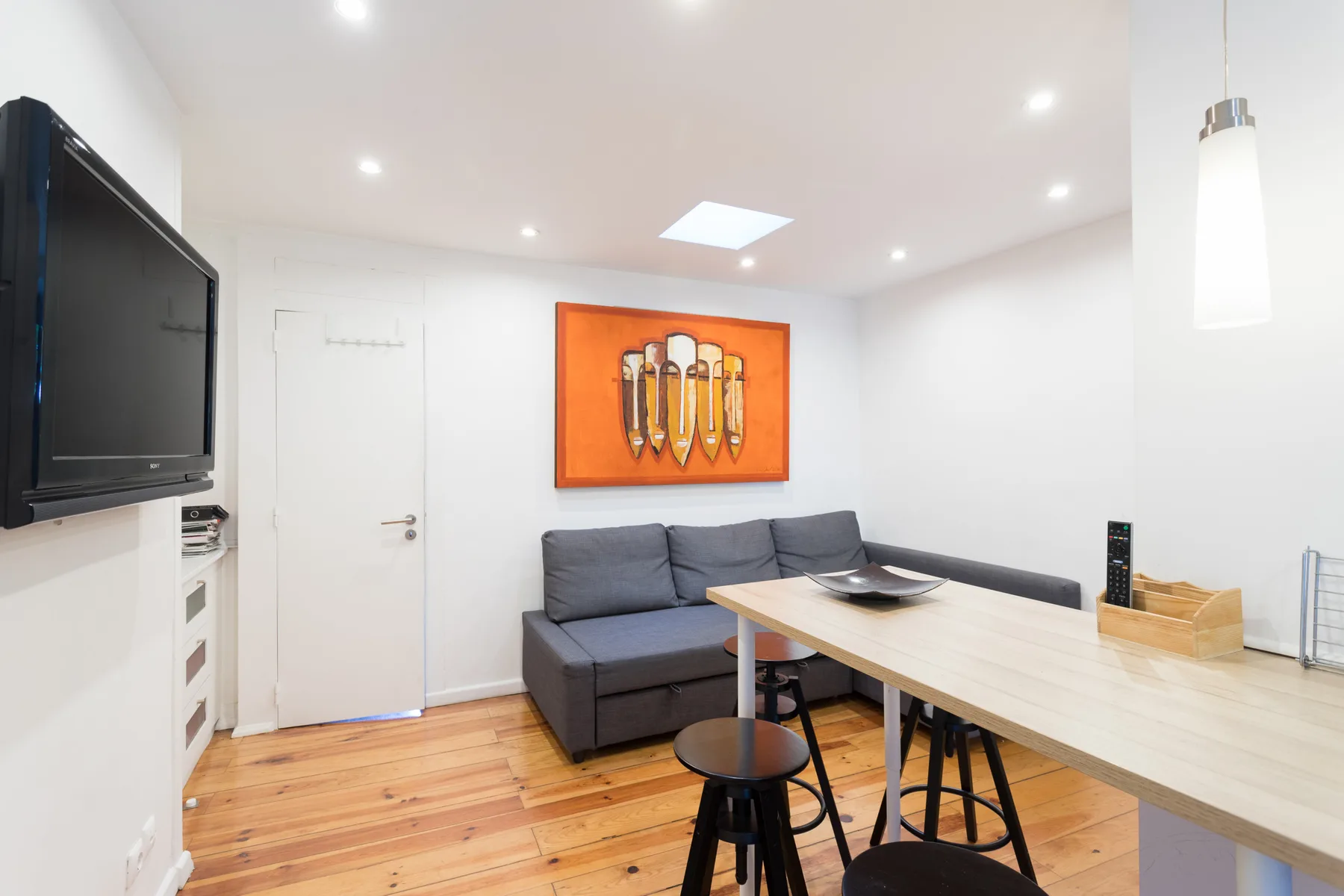 Un salón luminoso con asientos grises, una mesa de comedor y un televisor en la pared.