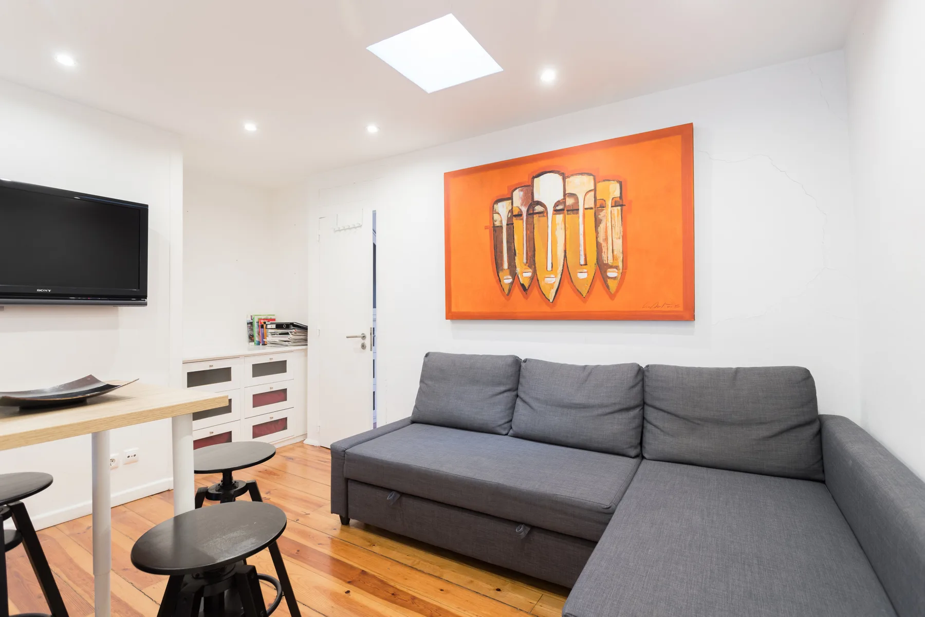 Una sala de estar con asientos grises, un televisor en la pared y arte naranja.