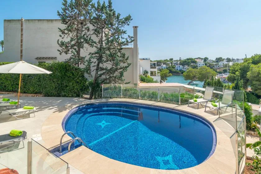 A swimming pool sits on a sunlit terrace with lounge chairs and a view of nearby buildings.
