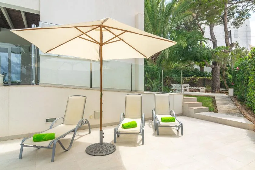 Un patio soleado con asientos reclinables, una sombrilla grande y escalones junto a la vegetación.