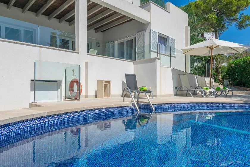 Una piscina junto a un edificio blanco moderno, con asientos reclinables y una sombrilla.