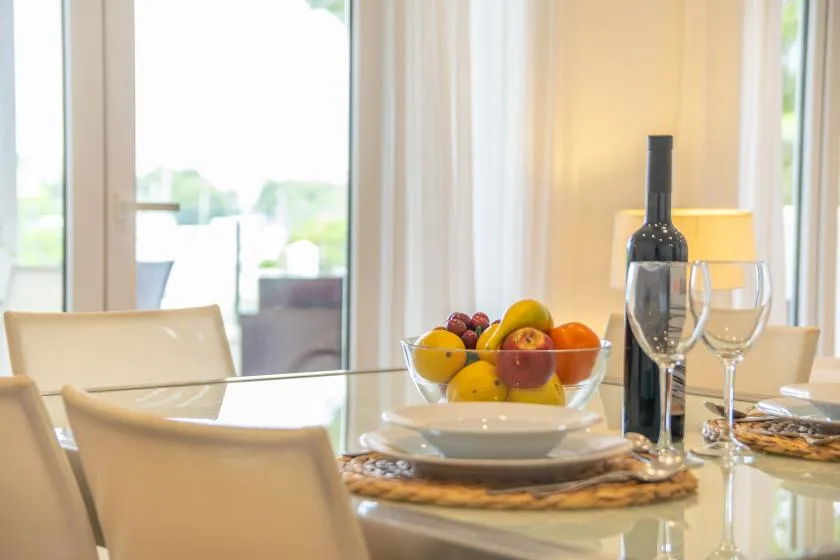 Un comedor luminoso con una mesa de cristal servida con fruta, vino y copas.