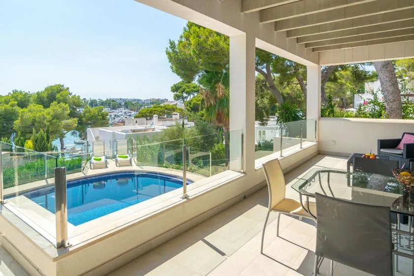 Una terraza cubierta con una mesa de cristal tiene vistas a una piscina y árboles verdes.
