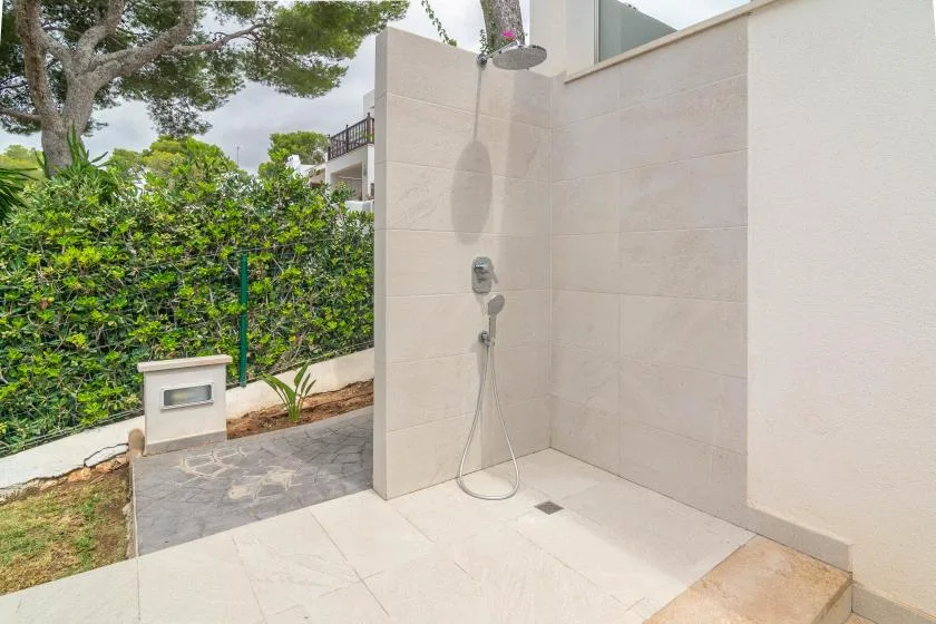 Una ducha exterior está junto a un seto frondoso y un patio pavimentado.