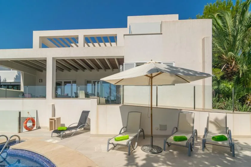 Un edificio blanco de estilo moderno da a una piscina, con asientos reclinables y una sombrilla.