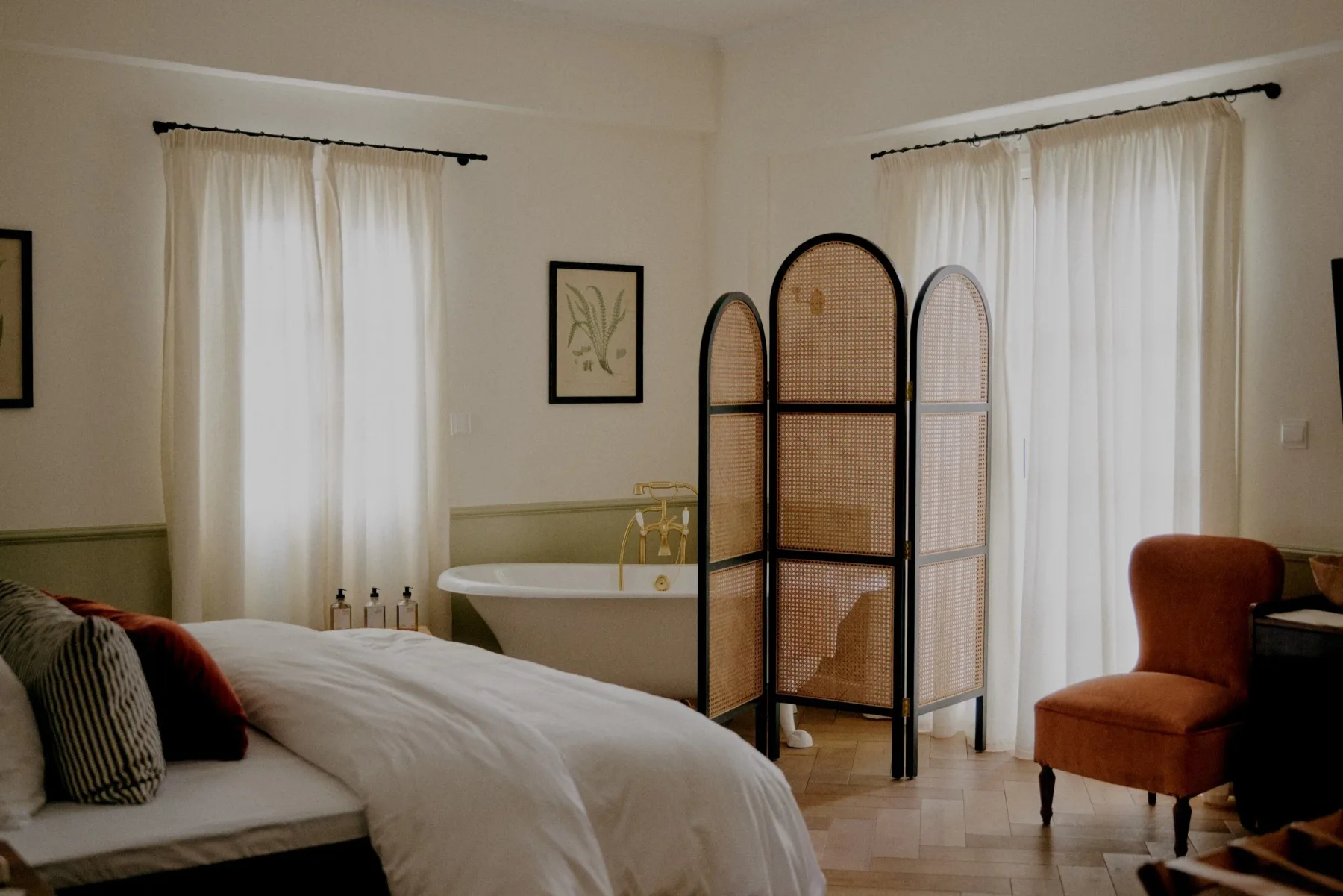 Un dormitorio con una cama, una bañera exenta, un biombo decorativo y una silla de color naranja quemado junto a una ventana.