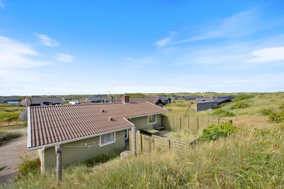 A low building with a sloped roof sits among grassy dunes under a clear sky.