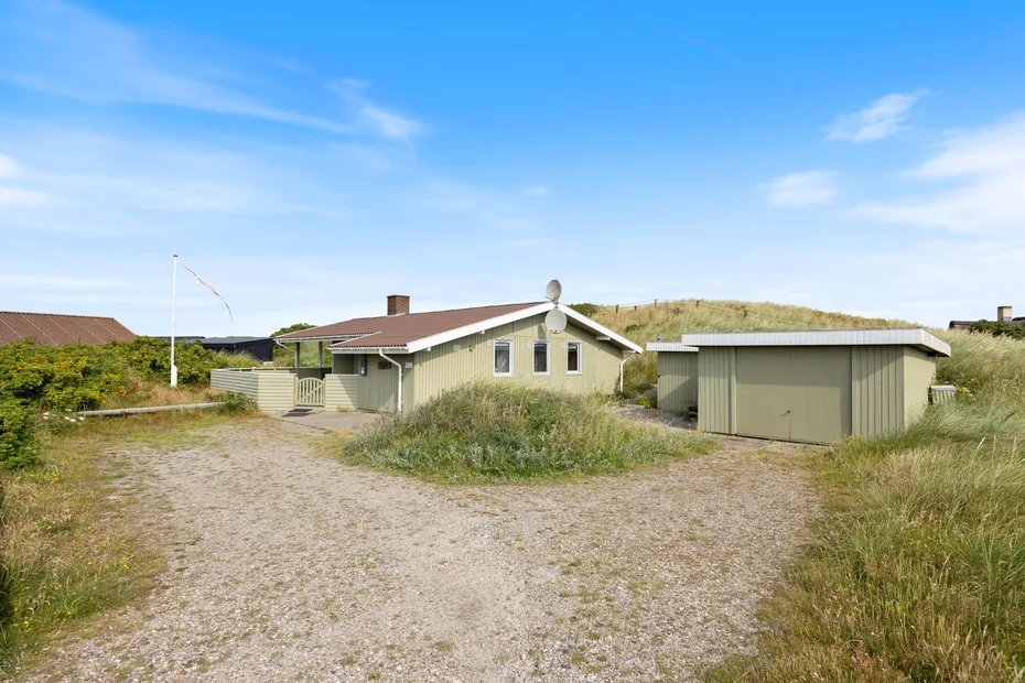 A small house and detached outbuilding sit beside a gravel drive amid grassy dunes.