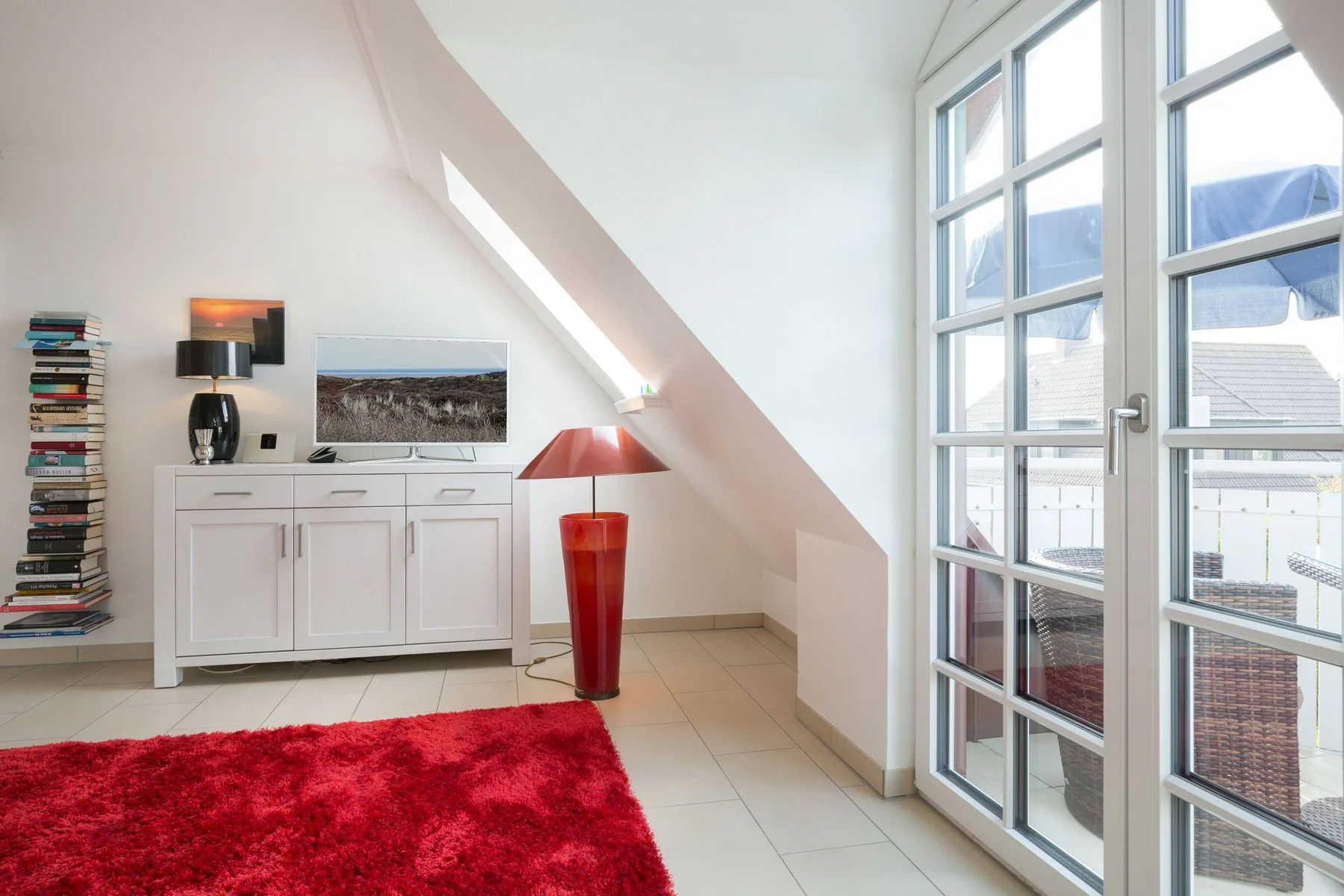 Una luminosa sala abuhardillada con muebles blancos, una alfombra roja y grandes puertas de cristal.
