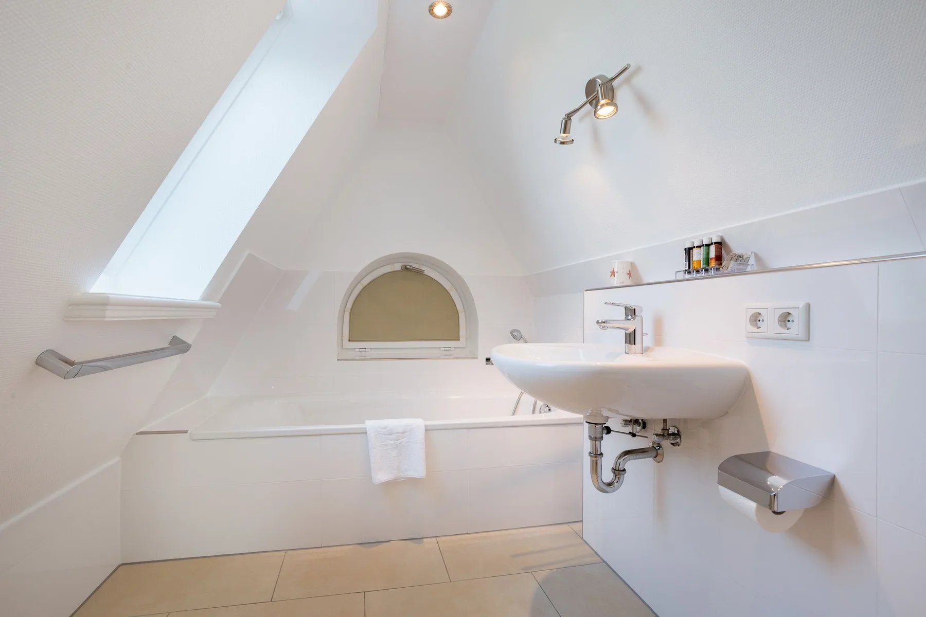Un baño luminoso con bañera y lavabo de pared bajo un techo inclinado.