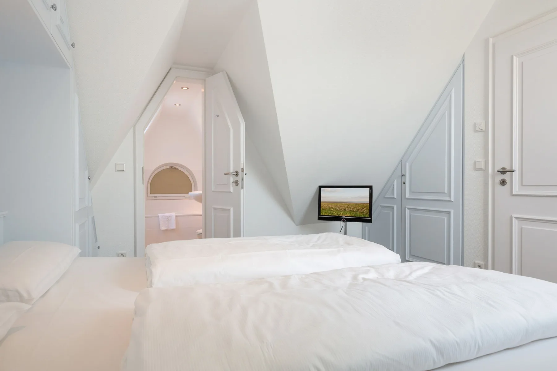Un dormitorio luminoso con ropa de cama blanca bajo un techo inclinado y una pantalla pequeña.